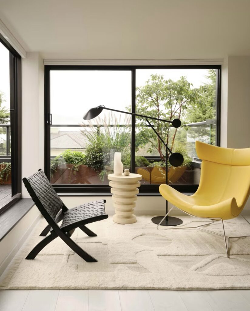A Bright London House Sunroom Features Modern Design: Yellow Armchair, Black Woven Chair, Unique Lamp, And Textured Rug. Large Windows Overlook A Lush Terrace.