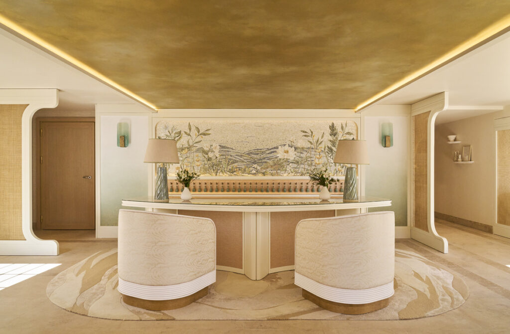 Elegant Boutique Hotel Reception Area With A Curved Cream And Beige Front Desk And Two Upholstered Armchairs In A Matching Palette. The Space Features A Large Botanical Mural In Soft Pastel Tones Behind The Counter, Flanked By Two Sculptural Table Lamps With Pleated Shades. The Ceiling Has A Luxurious Gold Finish With Recessed Linear Lighting That Adds Warmth. Walls Are Adorned With Minimalist Sconces And Architectural Details In White And Natural Textures, While A Patterned Neutral Rug Complements The Refined, Sophisticated Interior Design.