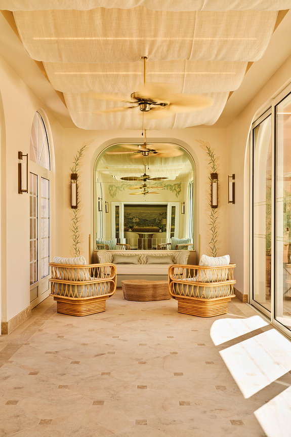 Serene Indoor Seating Nook With Two Rattan Armchairs And Plush Cushions, Positioned In Front Of A Large Decorative Mirror Framed By Hand-Painted Botanical Details, With Arched Windows, Soft Ceiling Drapes, And Classic Ceiling Fans Adding A Mediterranean Charm.