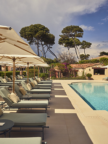 Luxurious Outdoor Pool Area Lined With Elegant Sun Loungers And Neutral-Toned Parasols, Surrounded By Lush Greenery And Tall Mediterranean Pine Trees Under A Partly Cloudy Sky, Creating A Relaxing Boutique Hotel Atmosphere.