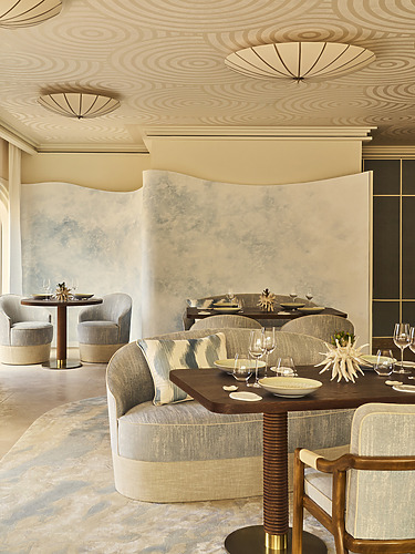 Elegant Dining Area With Curved Banquette Seating Upholstered In Neutral Tones And Soft Blue Accents, Round Wooden Tables Set With Fine Dining Tableware, And A Wave-Textured Wall Divider In Pastel Blue, Under Sculptural Ceiling Lights With Concentric Patterns.
