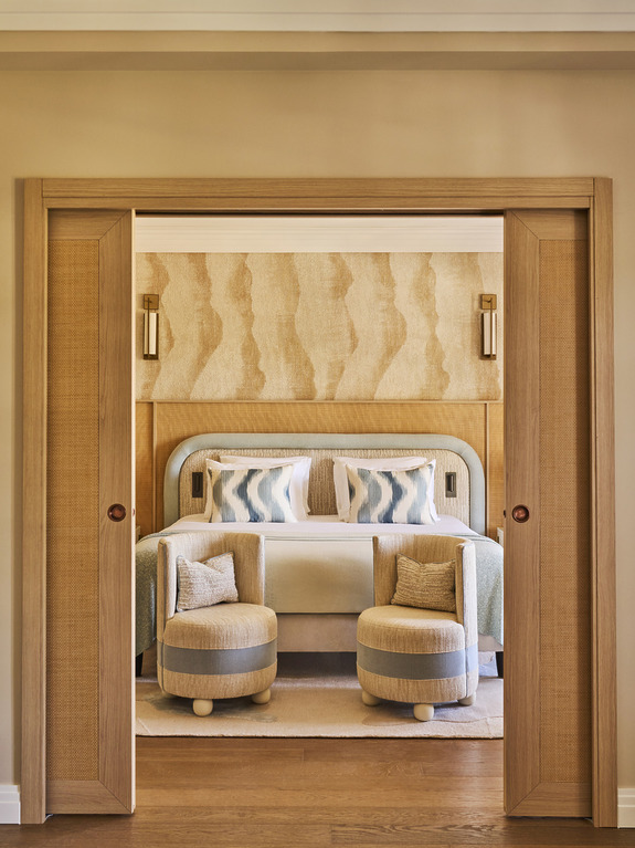 Elegant Boutique Hotel Bedroom With Sliding Wooden Doors Opening To Reveal A Plush Bed Framed By A Wavy-Textured Beige Headboard. The Room Features Soft Pastel Bedding With Geometric Patterns, Two Rounded Upholstered Stools At The Foot Of The Bed, Warm Wood Flooring, And Subtle Wall Sconces, Creating A Sophisticated And Inviting Atmosphere.
