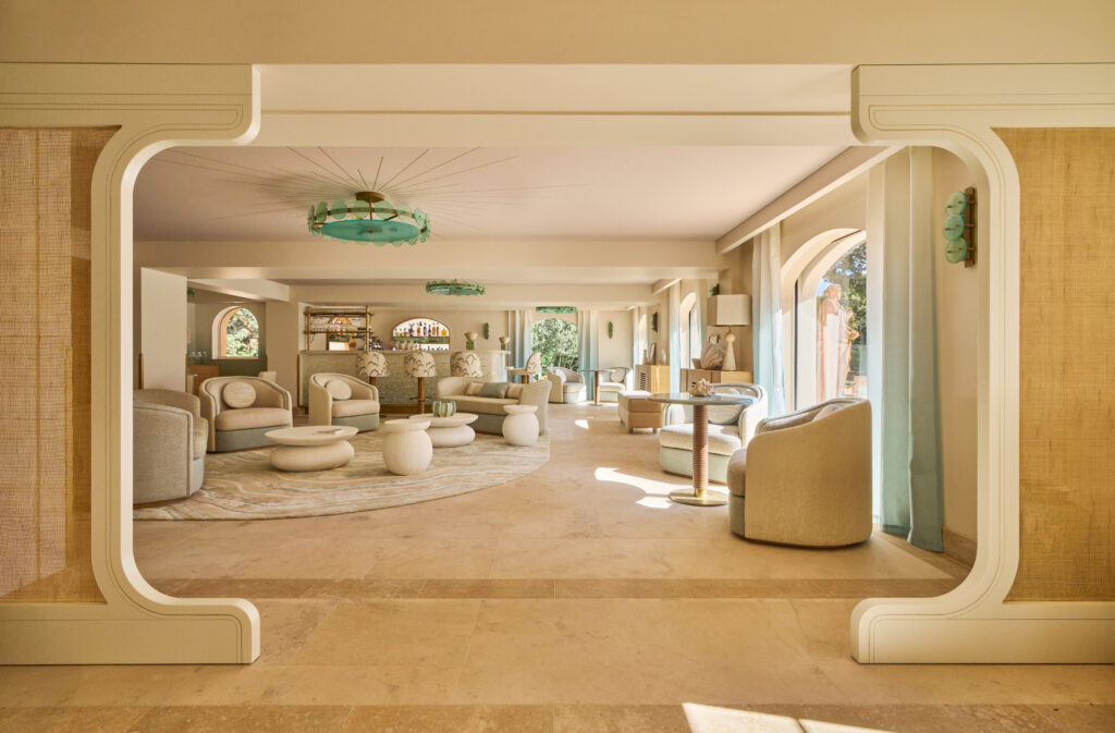 Boutique Hotel Lounge Interior Featuring A Spacious, Light-Filled Seating Area With Rounded Beige And Cream Sofas And Armchairs, Organic-Shaped White Coffee Tables, And A Large Neutral-Toned Rug. The Space Is Framed By Wide Arched Windows With Sheer Light-Blue Curtains, Allowing Abundant Natural Light. Architectural Details Include Art Deco–Inspired White Moldings, Green Glass Ceiling Chandeliers, And Matching Wall Sconces. The Beige Stone Flooring Enhances The Elegant, Sophisticated, And Relaxed Mediterranean Design Aesthetic.