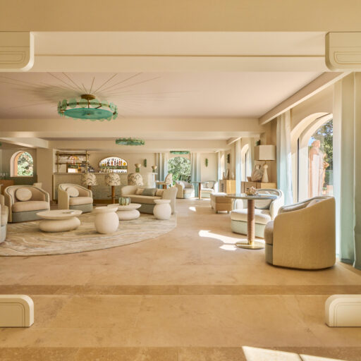 Boutique Hotel Lounge Interior Featuring A Spacious, Light-Filled Seating Area With Rounded Beige And Cream Sofas And Armchairs, Organic-Shaped White Coffee Tables, And A Large Neutral-Toned Rug. The Space Is Framed By Wide Arched Windows With Sheer Light-Blue Curtains, Allowing Abundant Natural Light. Architectural Details Include Art Deco–Inspired White Moldings, Green Glass Ceiling Chandeliers, And Matching Wall Sconces. The Beige Stone Flooring Enhances The Elegant, Sophisticated, And Relaxed Mediterranean Design Aesthetic.