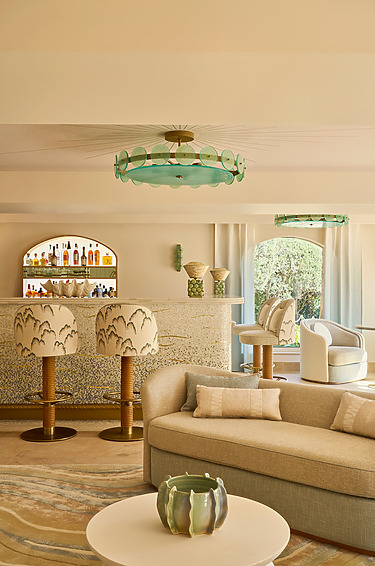 Stylish Lounge And Bar Area Featuring A Pale Beige Sofa With Cream Cushions, A Round White Coffee Table With Decorative Ceramics, A Chic Bar With Scalloped-Edge Stools And A Mirrored Back Shelf, All Beneath A Statement Green Glass Ceiling Light And Arched Curtains Opening To A Garden View.
