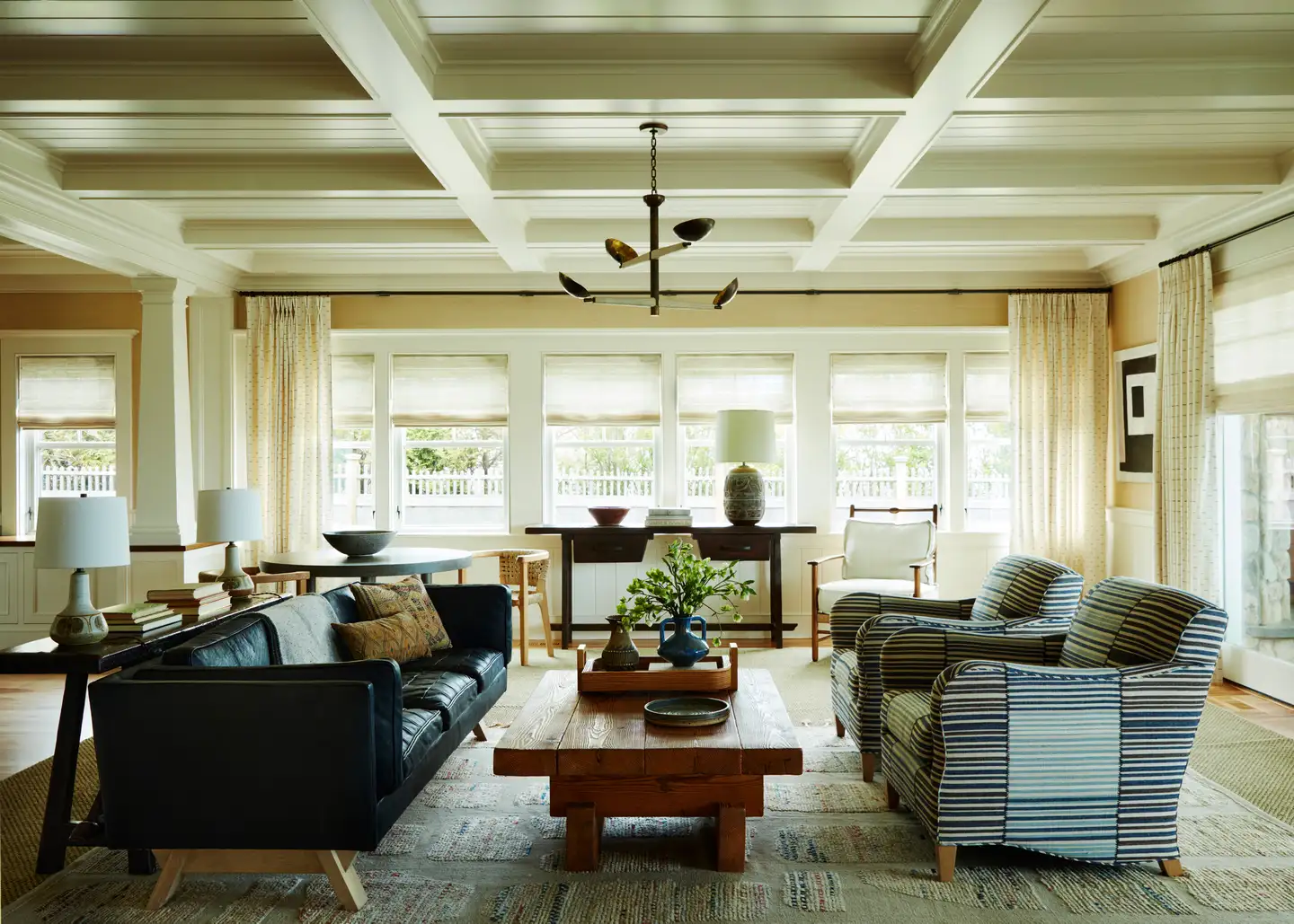 This Is A Living Room With A Black Leather Sofa, Two Striped Armchairs, And A Wooden Coffee Table. The Room Has A High Ceiling With Exposed Beams, And There Are Several Windows That Let In Natural Light. There Is Also A Dining Table With Chairs In The Background. The Room Features A Green Rugs That Adds A Pop Of Color To The Space.