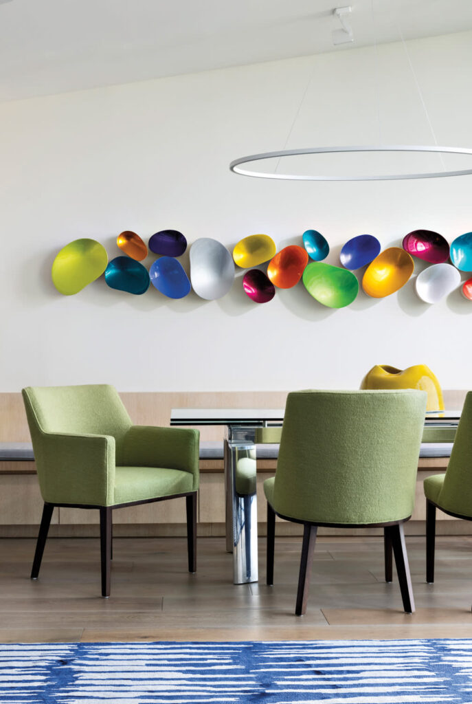 A Stylish Dining Space Featuring Light Green Upholstered Chairs Around A Sleek Glass Dining Table With Chrome Legs. The Focal Point Is A Vibrant Wall-Mounted Sculpture Composed Of Multi-Colored, Bowl-Like Forms Arranged In A Flowing Pattern. The Neutral Wall And Wooden Flooring Highlight The Artwork's Bold Palette, Adding Artistic Flair To The Minimalist Setting.

