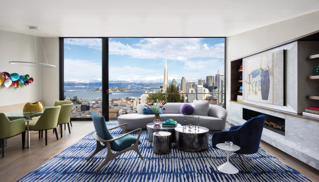 A Modern Living Room And Dining Area With Floor-To-Ceiling Windows Offering A Stunning Panoramic View Of San Francisco, Including The Transamerica Pyramid And The Bay. The Interior Features Contemporary Furniture, Including A Curved Light-Gray Sofa, Blue And Green Accent Chairs, A Blue-Patterned Rug, And Artistic Decor. The Dining Area Includes Green Upholstered Chairs And A Glass Table, Complemented By A Colorful Wall Sculpture. The Space Is Decorated With Vibrant Artwork And Modern Lighting, Creating A Stylish And Inviting Atmosphere With A Modern Area Rugs.