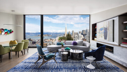 A Modern Living Room And Dining Area With Floor-To-Ceiling Windows Offering A Stunning Panoramic View Of San Francisco, Including The Transamerica Pyramid And The Bay. The Interior Features Contemporary Furniture, Including A Curved Light-Gray Sofa, Blue And Green Accent Chairs, A Blue-Patterned Rug, And Artistic Decor. The Dining Area Includes Green Upholstered Chairs And A Glass Table, Complemented By A Colorful Wall Sculpture. The Space Is Decorated With Vibrant Artwork And Modern Lighting, Creating A Stylish And Inviting Atmosphere With A Modern Area Rugs.