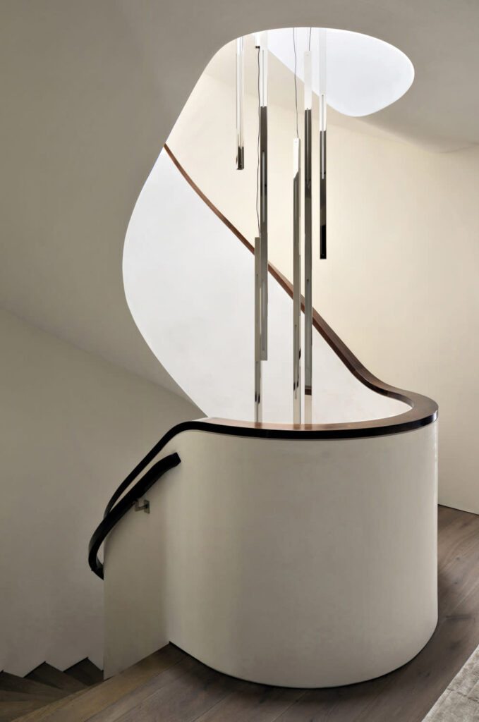 A Sculptural, Curved Staircase Featuring Sleek White Plaster Walls And A Dark Wood Handrail, Blending Seamlessly Into Minimalist Architecture. Suspended From Above, An Array Of Slender Metallic Pendant Lights Adds A Contemporary, Artistic Touch To The Space. The Soft Natural Light And Smooth Textures Create A Tranquil, Gallery-Like Ambiance.

