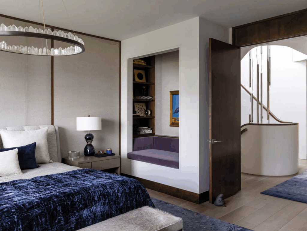 A Sophisticated Modern Bedroom Features A Plush Upholstered Bed Adorned With Navy And Cream Pillows And A Rich Navy Velvet Bedspread. A Sculptural Black Lamp Sits On The Sleek Bedside Table Beneath A Striking Circular Chandelier With Crystal-Like Elements. A Built-In Reading Nook With A Purple Cushion And Surrounding Shelves Is Inset Into The Wall, Adding Depth And Function. Through An Open Door, A Softly Curved Staircase And Hanging Pendant Lights Are Visible, Unifying The Home's Fluid, Architectural Aesthetic.