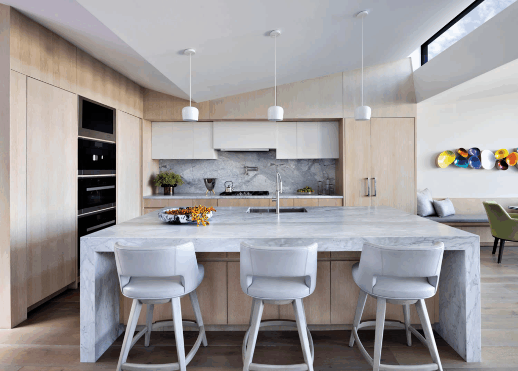 A Refined Modern Kitchen Showcases Light-Toned Wood Cabinetry And A Dramatic Marble Island With Waterfall Edges, Accompanied By Three Upholstered Bar Stools In Soft Gray. Minimalist White Pendant Lights Hang Above The Island, While The Marble Backsplash And Integrated Appliances Enhance The Streamlined Aesthetic. To The Right, A Cozy Banquette Seating Area And Lime-Green Dining Chairs Add Warmth And Color, Complemented By A Striking Wall Installation Of Multicolored Sculptural Forms.

