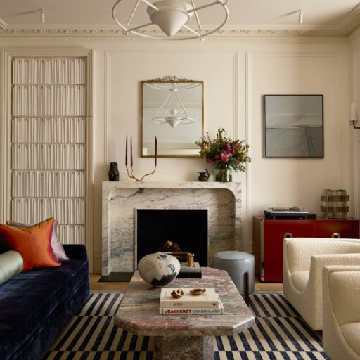 An Elegant Living Room With A Marble Fireplace, Blue Sofa, And White Armchairs. The Room Features A Unique Textured Door, A Vibrant Abstract Painting, And A Decorative Mirror. This Space Embodies Elevated Living With Its Artistic Flair And Sophisticated Design.
